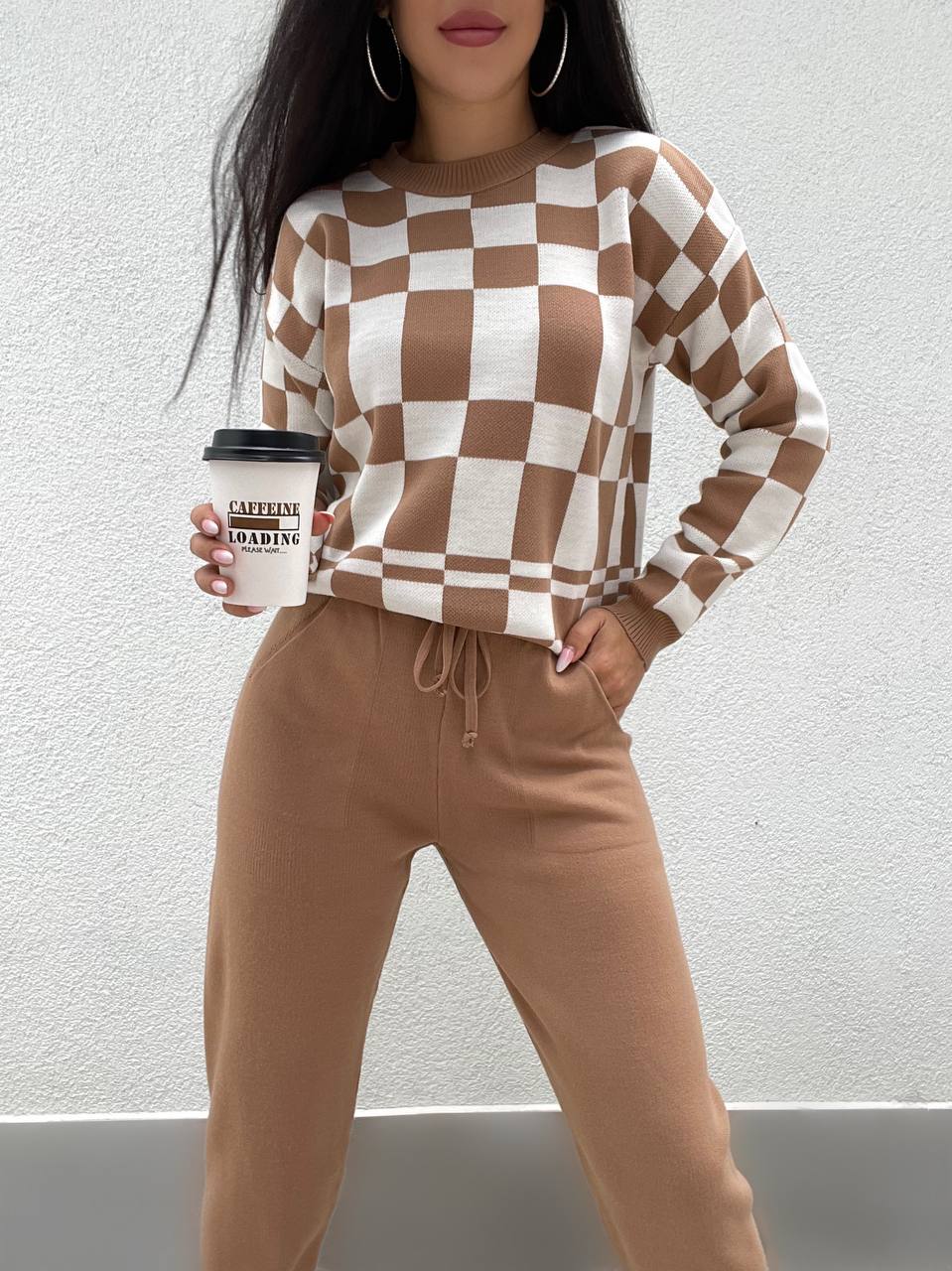 Person wearing a checkered brown and white sweater and beige pants, holding a coffee cup.