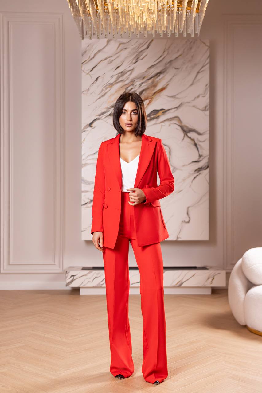 Woman in a red suit standing in an elegant room with marble wall and chandelier.