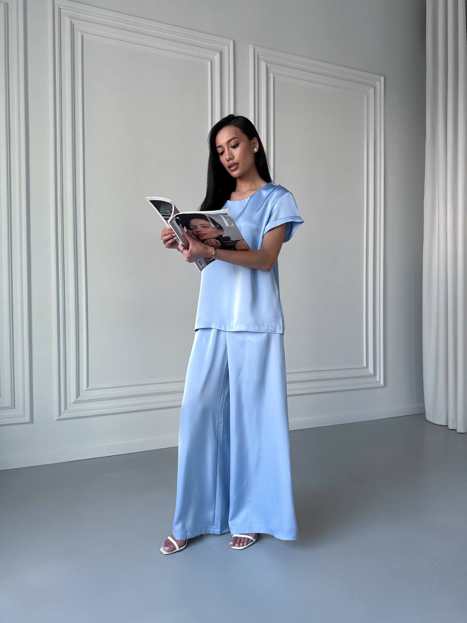 Woman in a light blue outfit reading a book against a white paneled wall.