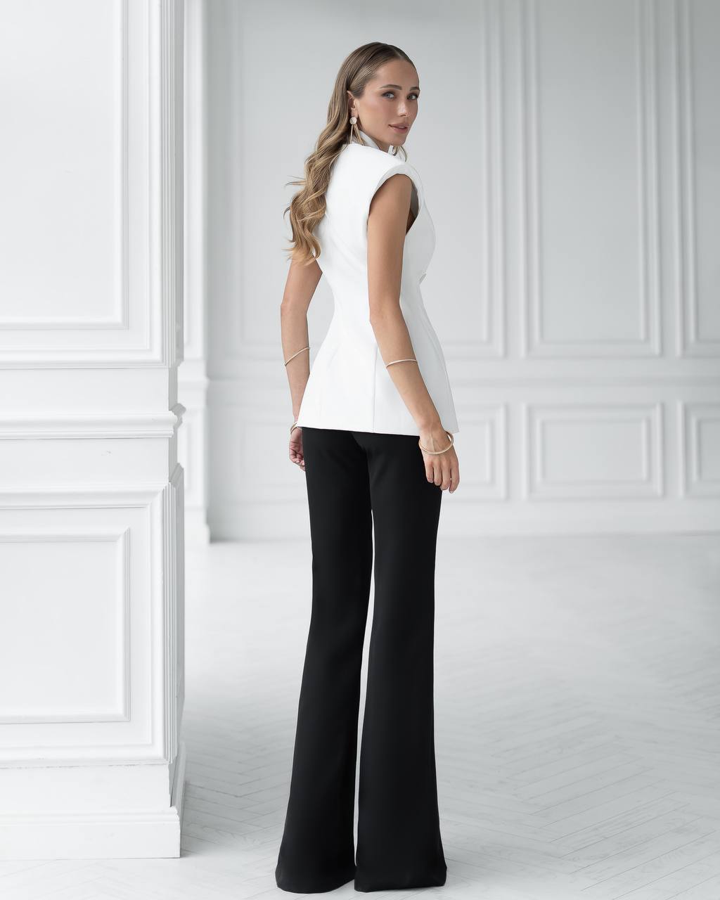 Woman wearing a white sleeveless top and black pants against a white paneled wall.