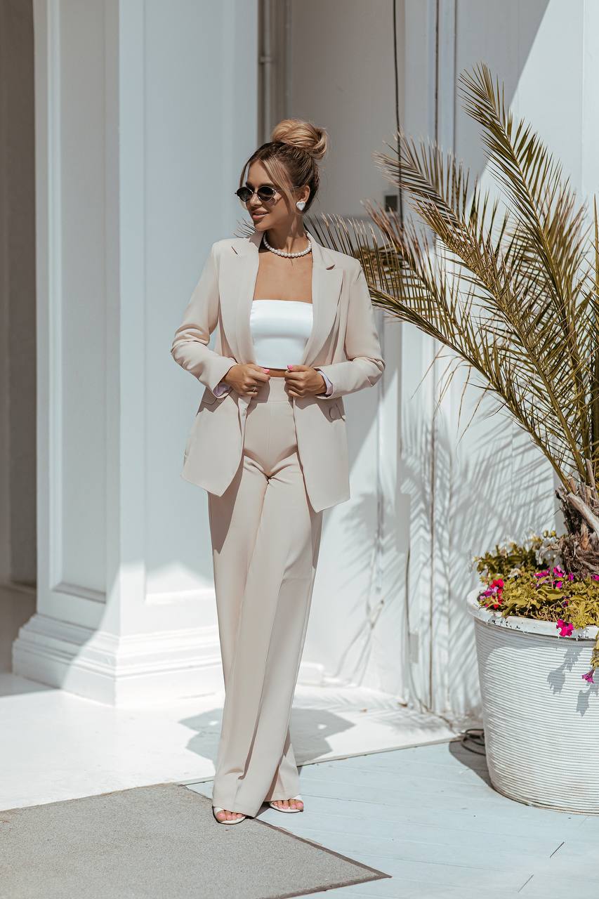 Woman in a light-colored suit standing outdoors with a palm tree and flowers in the background