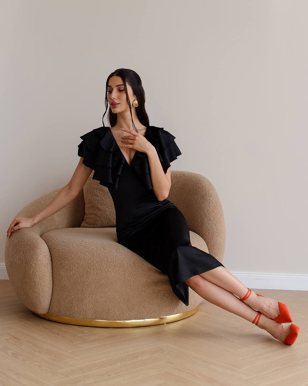 Woman in a black dress sitting on a beige armchair with a plain background