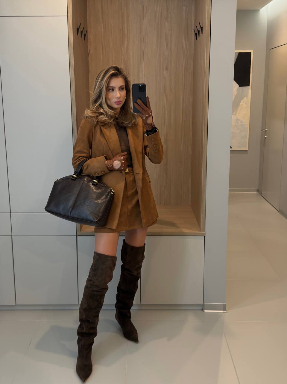 Woman taking a mirror selfie wearing a brown coat, skirt, and knee-high boots in a minimalistic room.