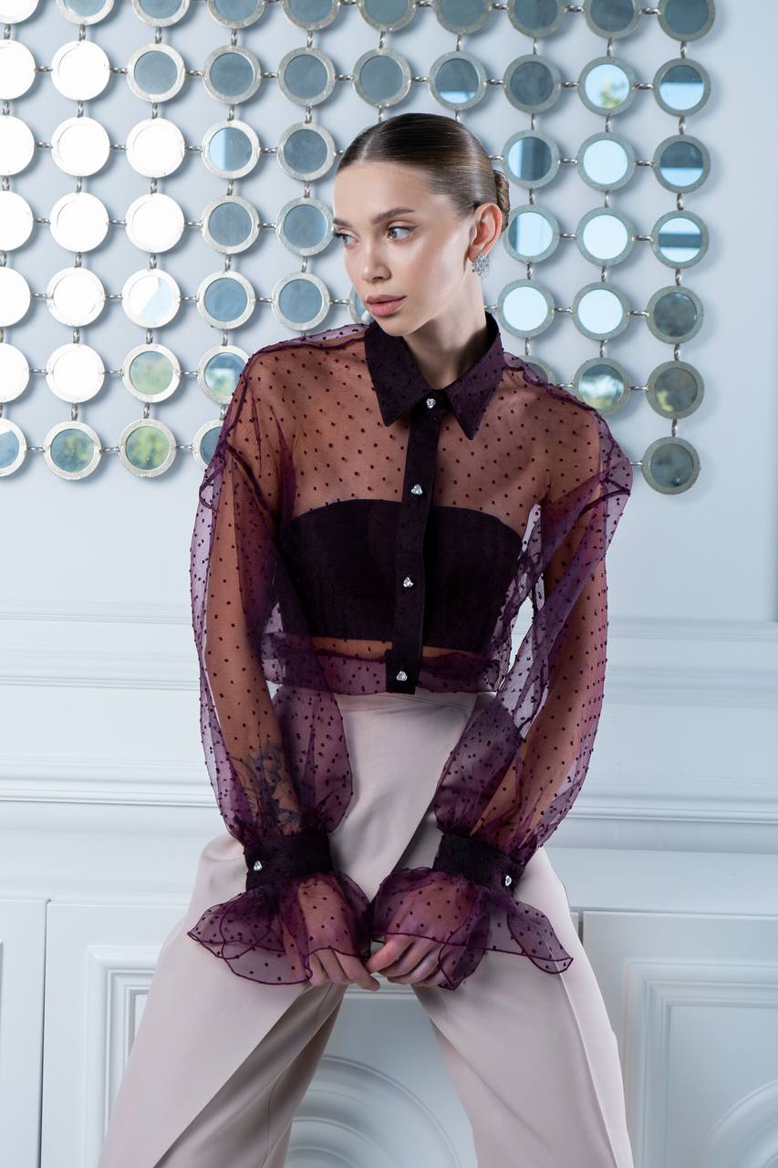 Woman wearing a sheer purple top over a black shirt with a decorative wall in the background