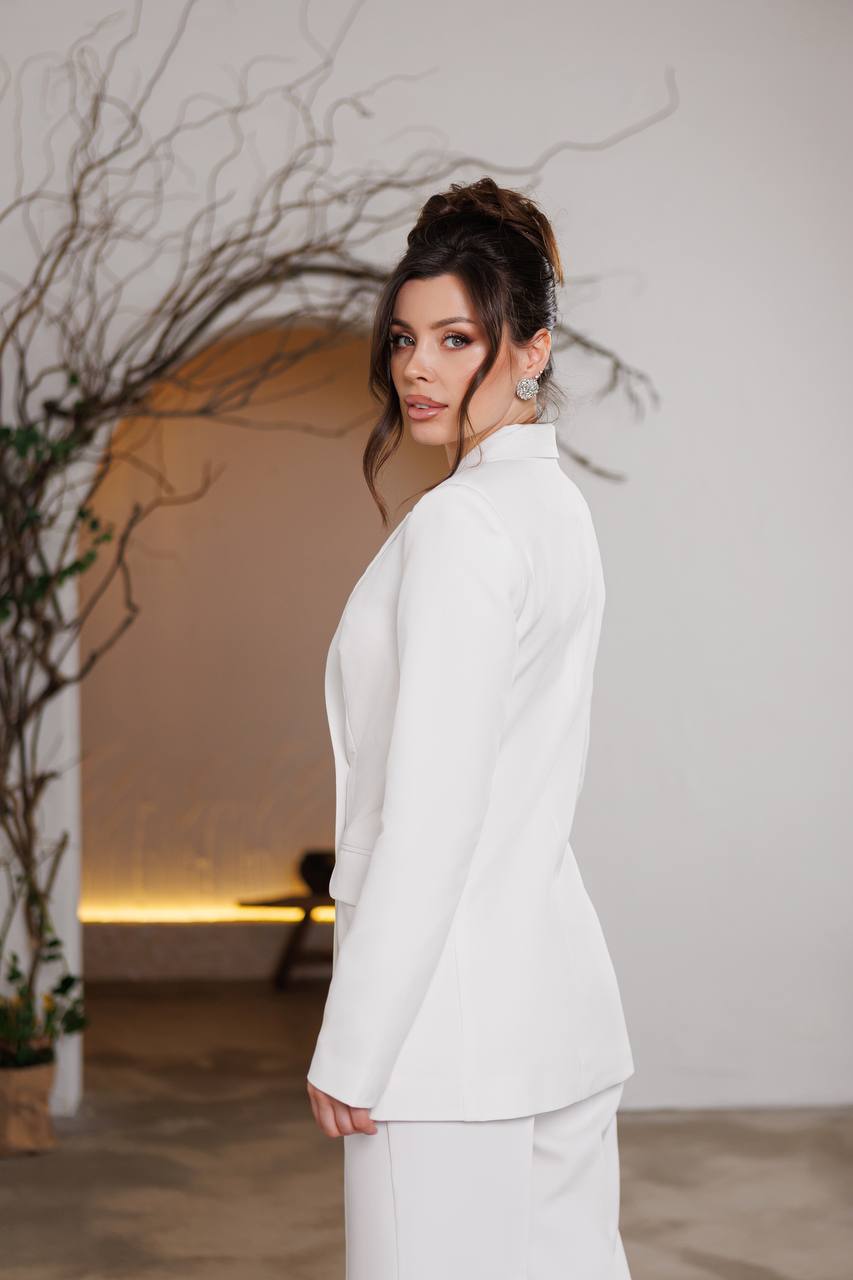 Woman in a white suit standing in a softly lit room with decorative plants.