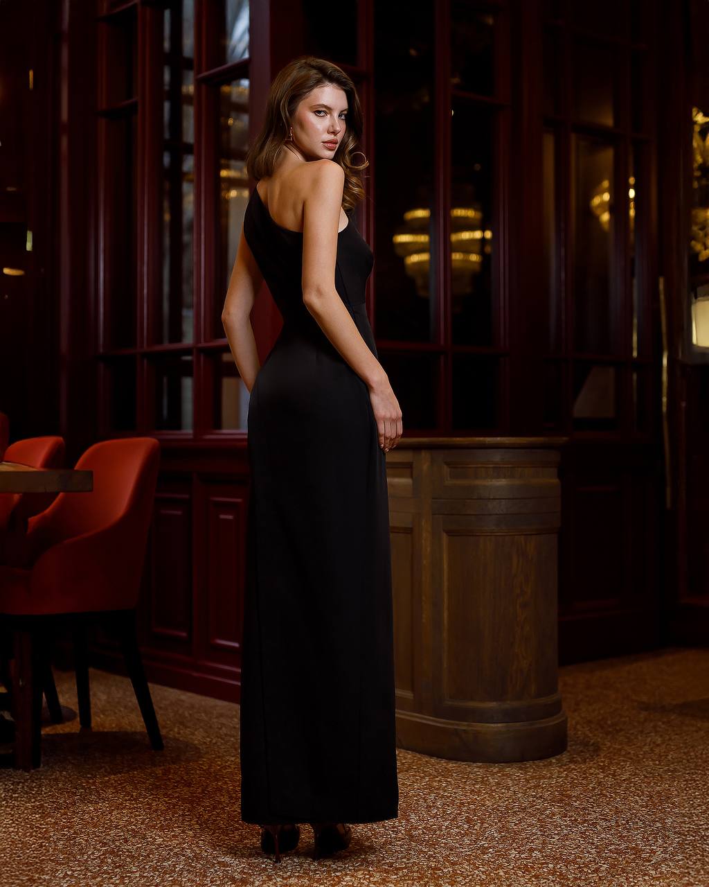 Woman in a black dress standing in an indoor setting with wooden elements and red chairs.
