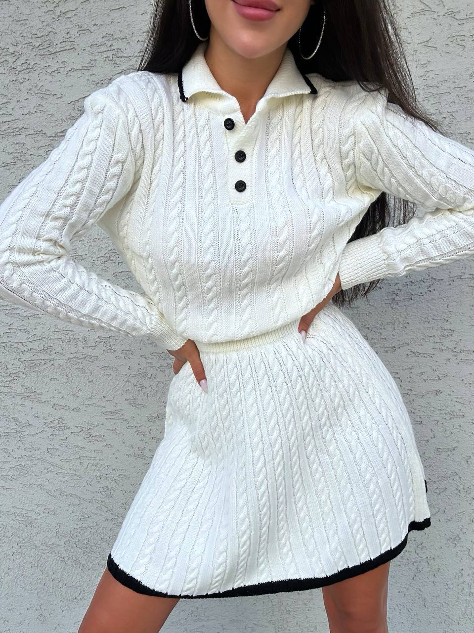Woman wearing a white cable knit outfit with black accents against a gray background