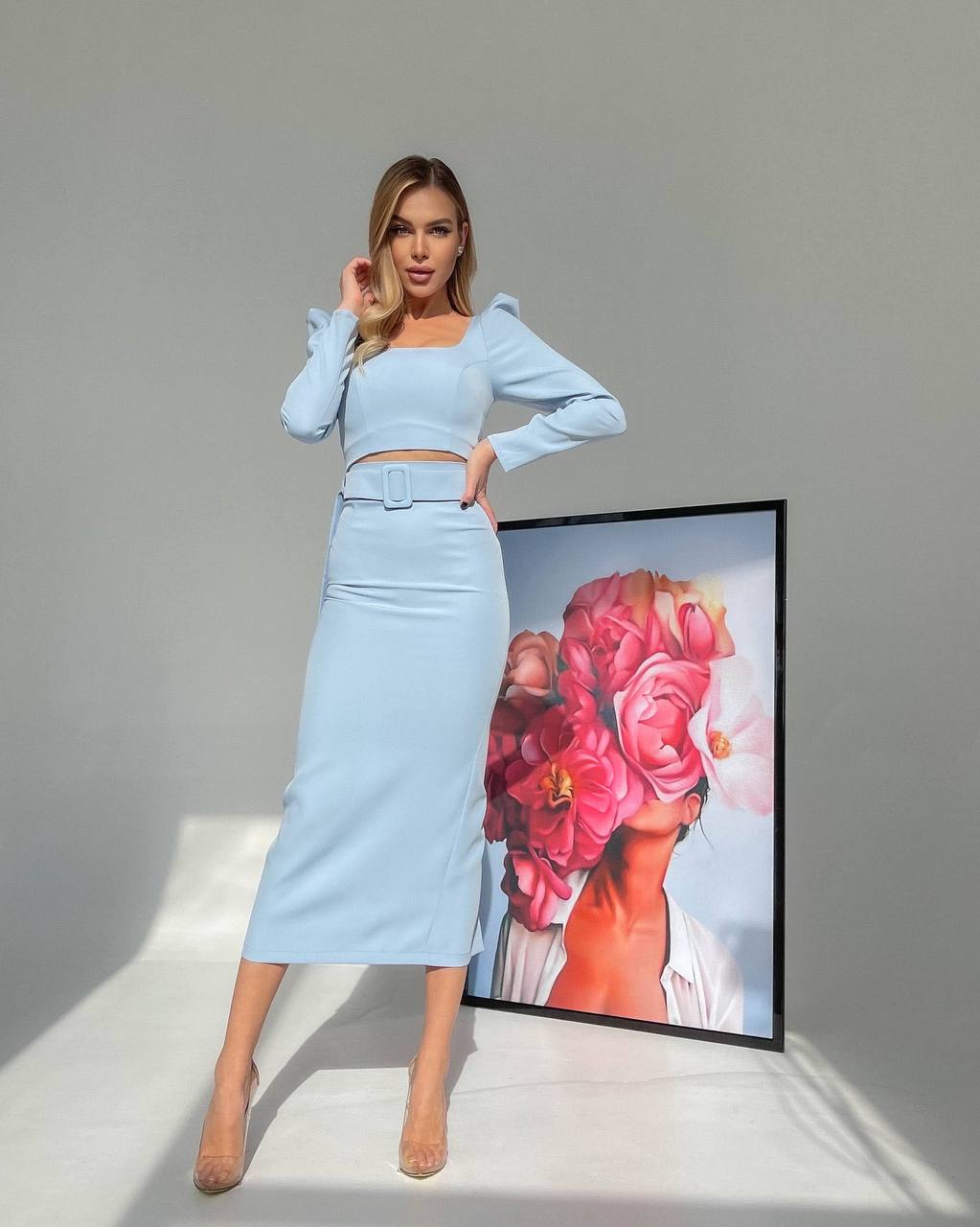 Woman in a light blue outfit standing next to a framed artwork on a gray background
