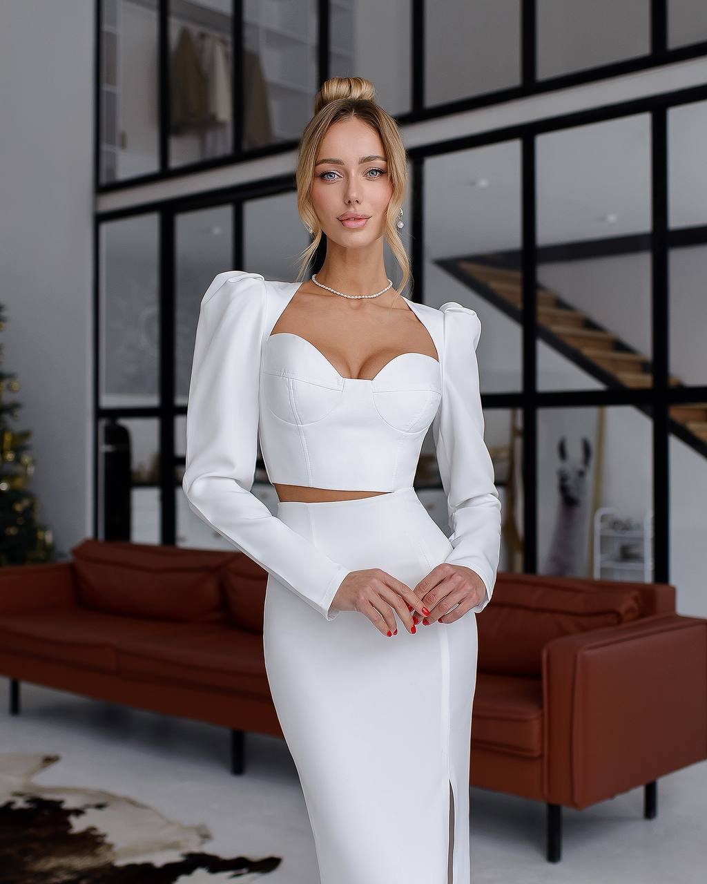 Woman in a white outfit standing in a modern indoor setting with a brown sofa and glass walls.