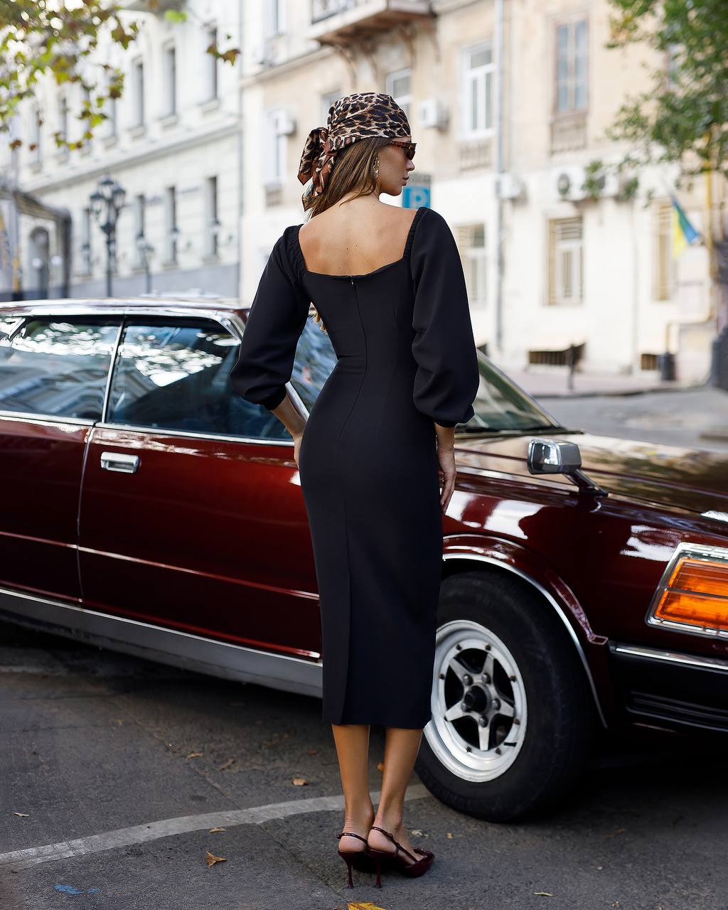 Woman in a black dress standing next to a vintage car on a city street.