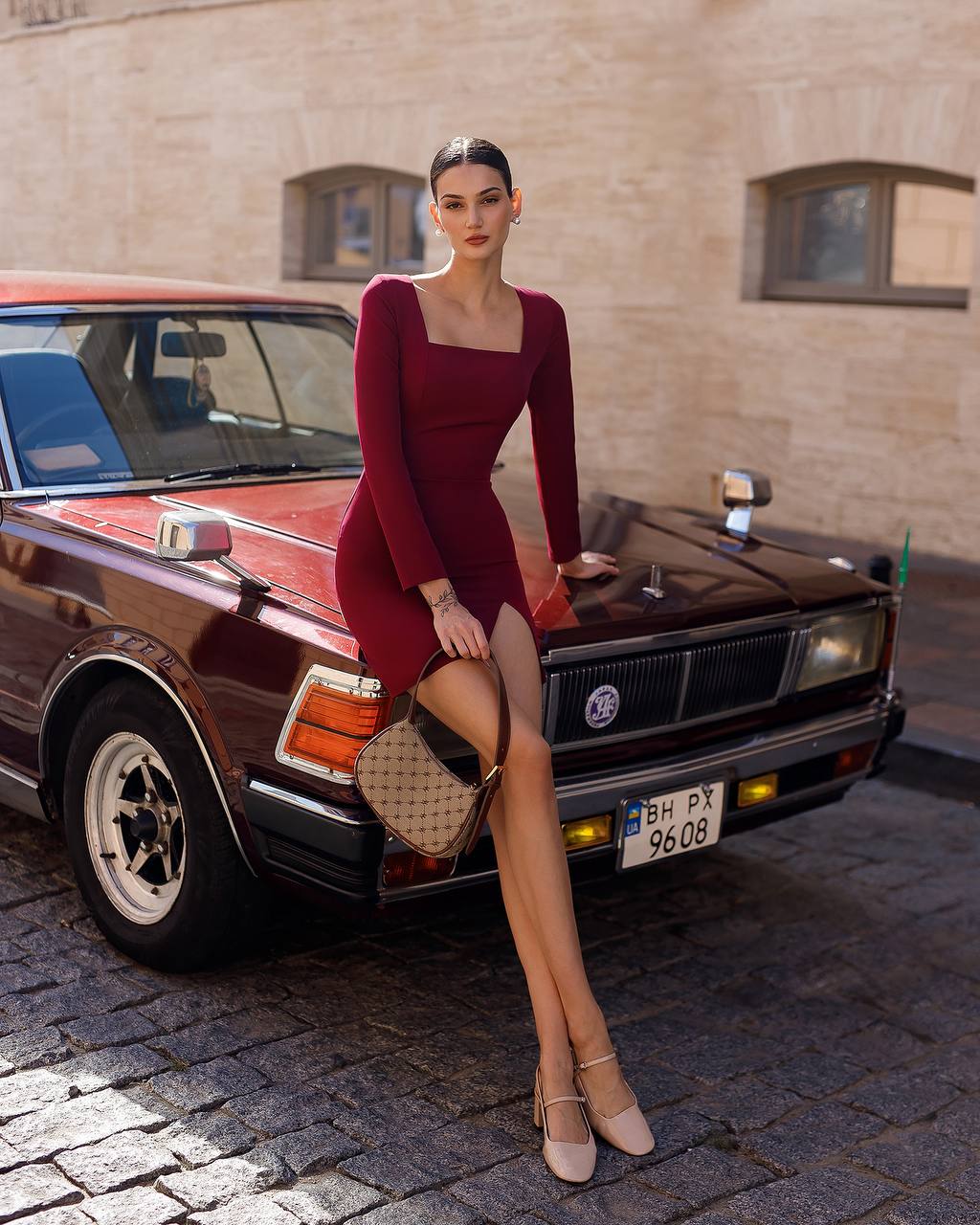 Woman in a red dress posing in front of a vintage car on a cobblestone street.