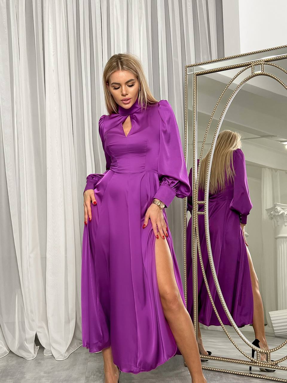 Woman in a purple dress standing in front of a mirror with white curtains in the background