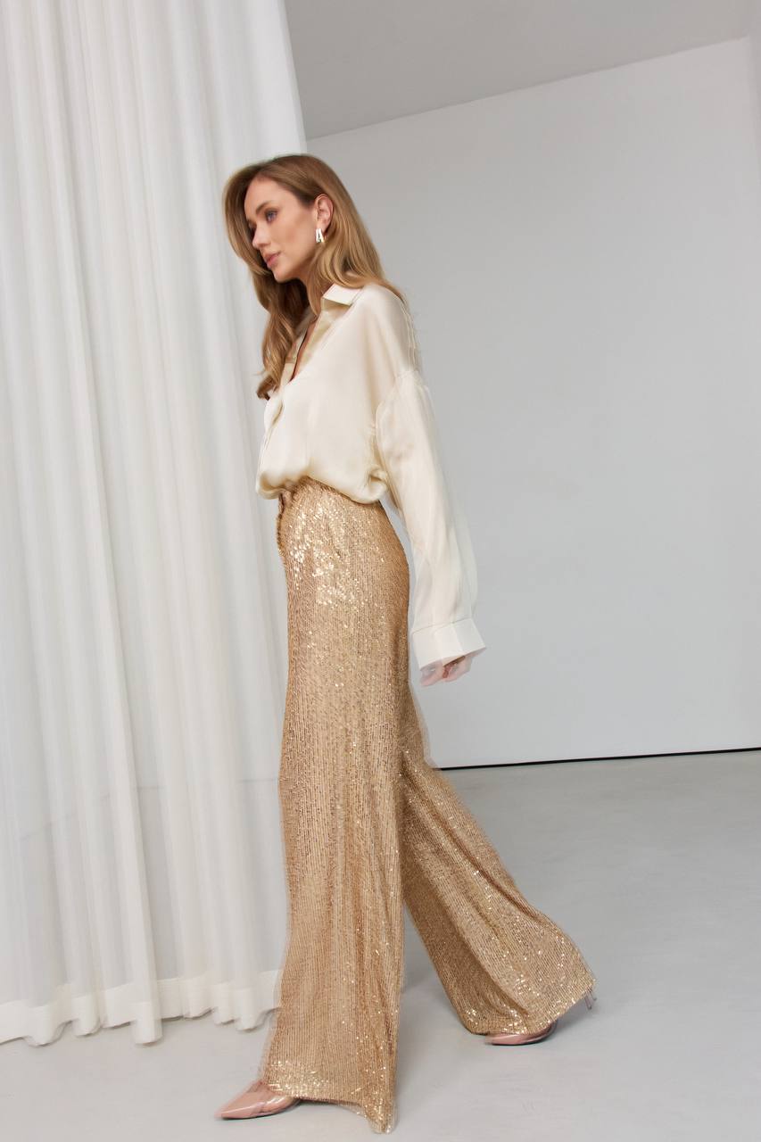 Woman wearing a white blouse and gold sequin pants against a white background