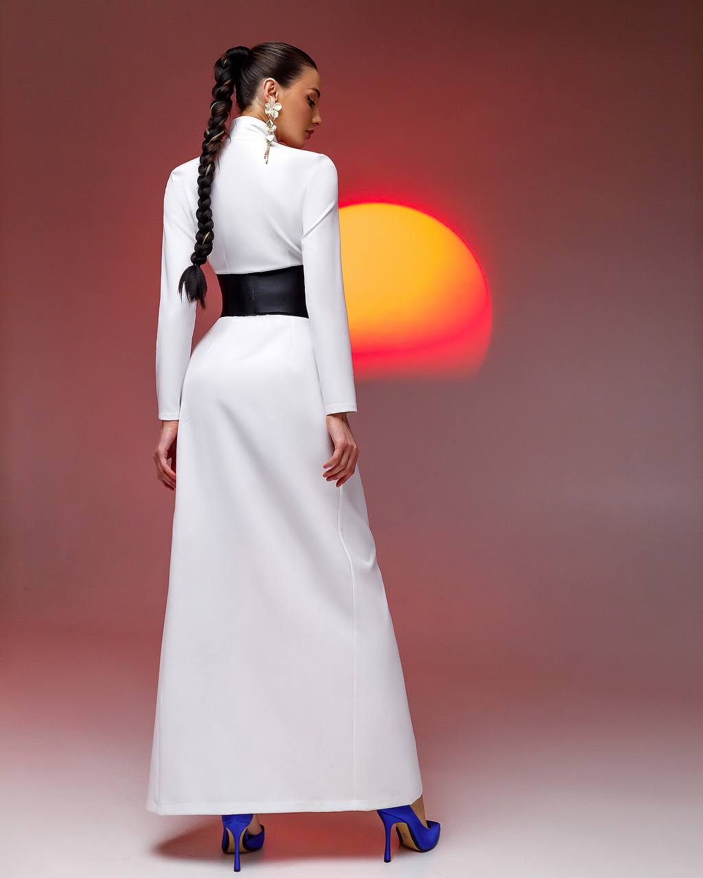 Woman in a white dress with a black belt standing against a sunset background