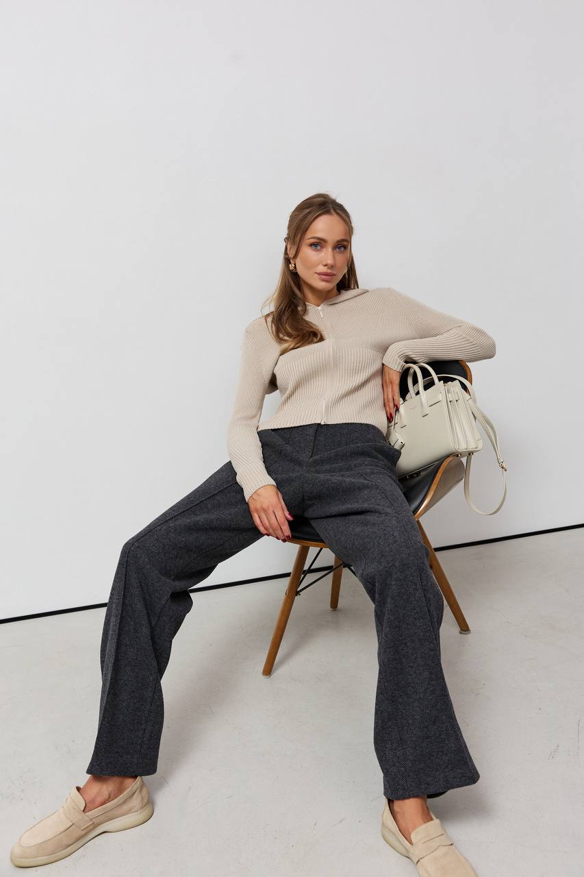 Woman sitting on a chair wearing a beige sweater and dark pants, holding a white handbag.