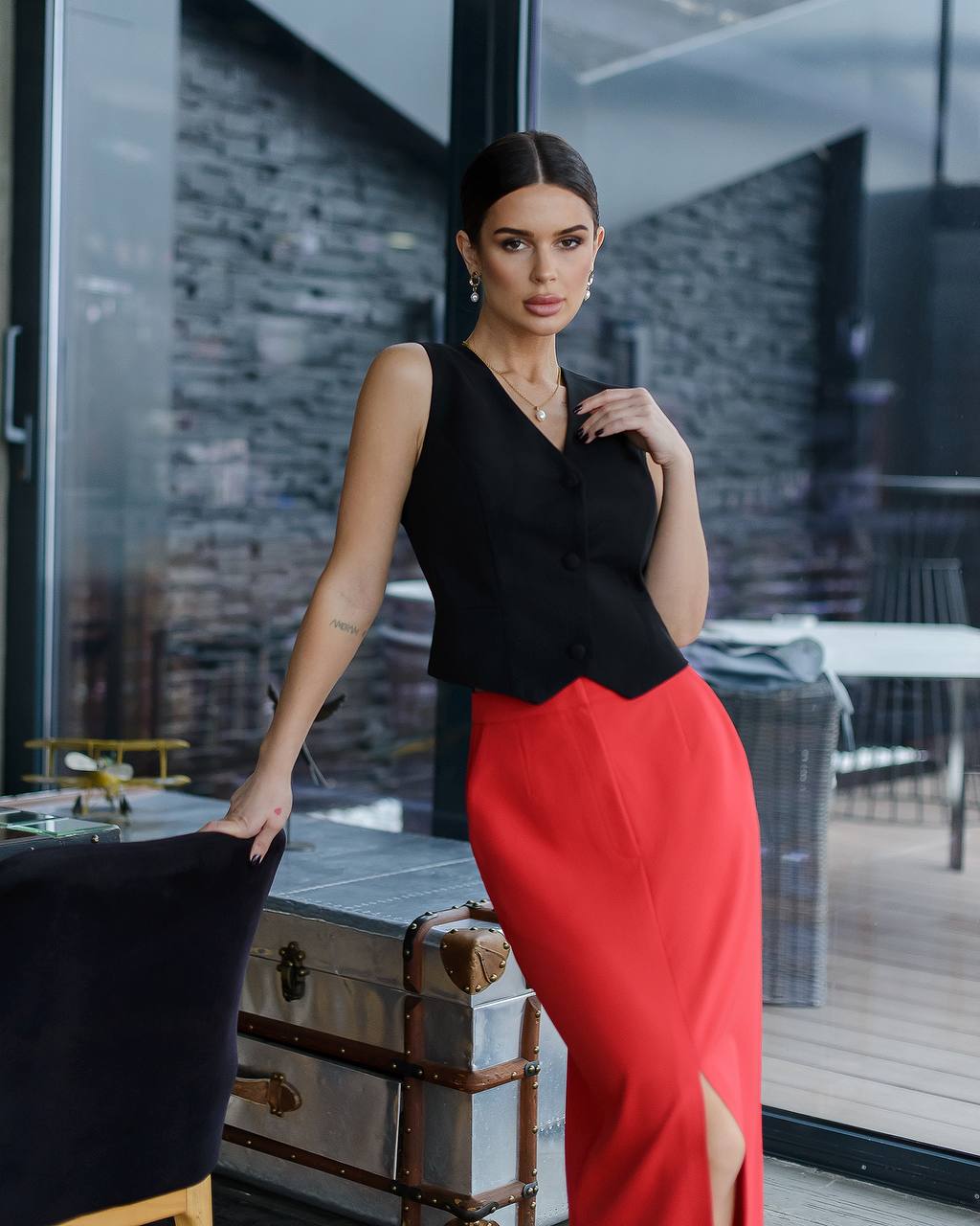 Woman in a black top and red skirt standing in an indoor setting with a cityscape view.