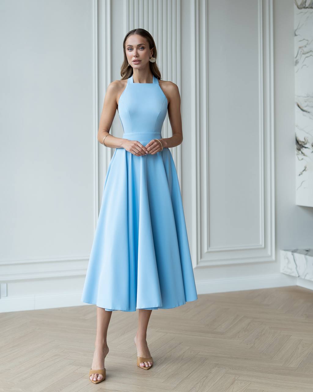 Woman wearing a light blue dress standing in an elegant room with white walls and wooden floor.