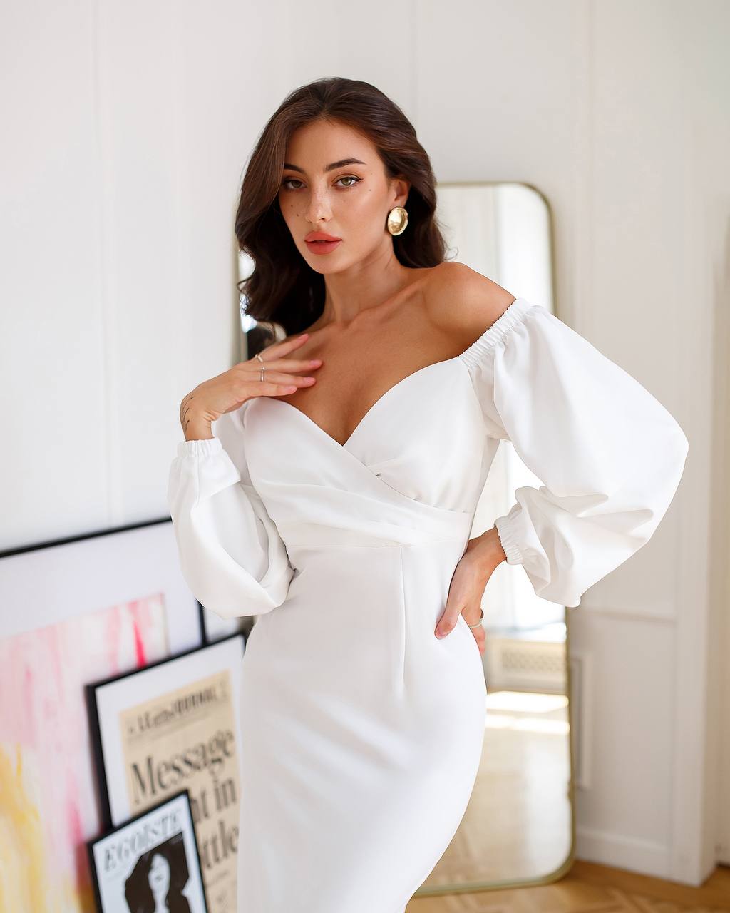 Woman wearing a white off-shoulder dress in a room with a mirror and decor.