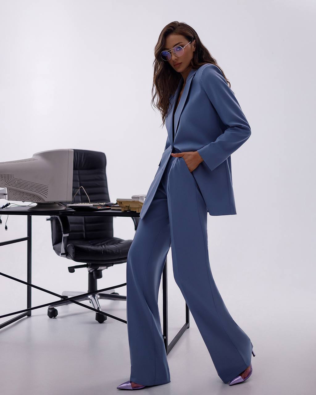 Woman in a blue suit standing in an office setting with a desk and chair.