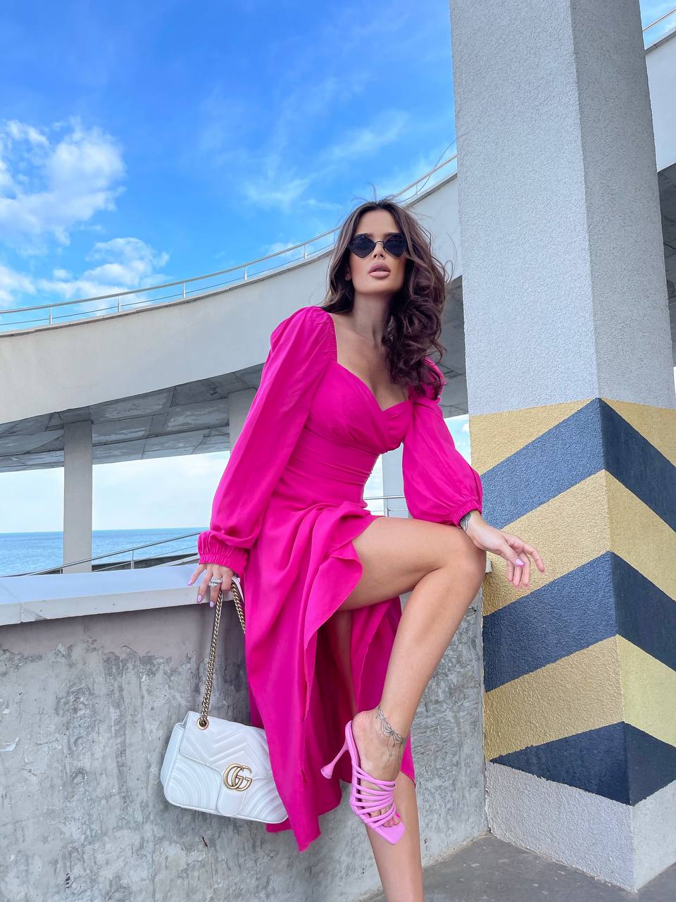 Woman in a bright pink dress standing against a geometric wall with ocean view.
