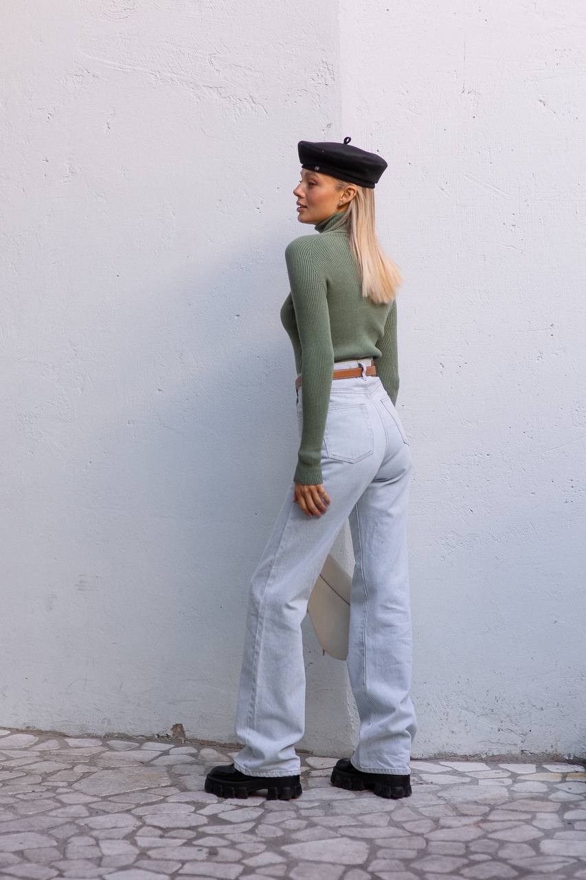 Person wearing a green sweater and white jeans against a white wall.