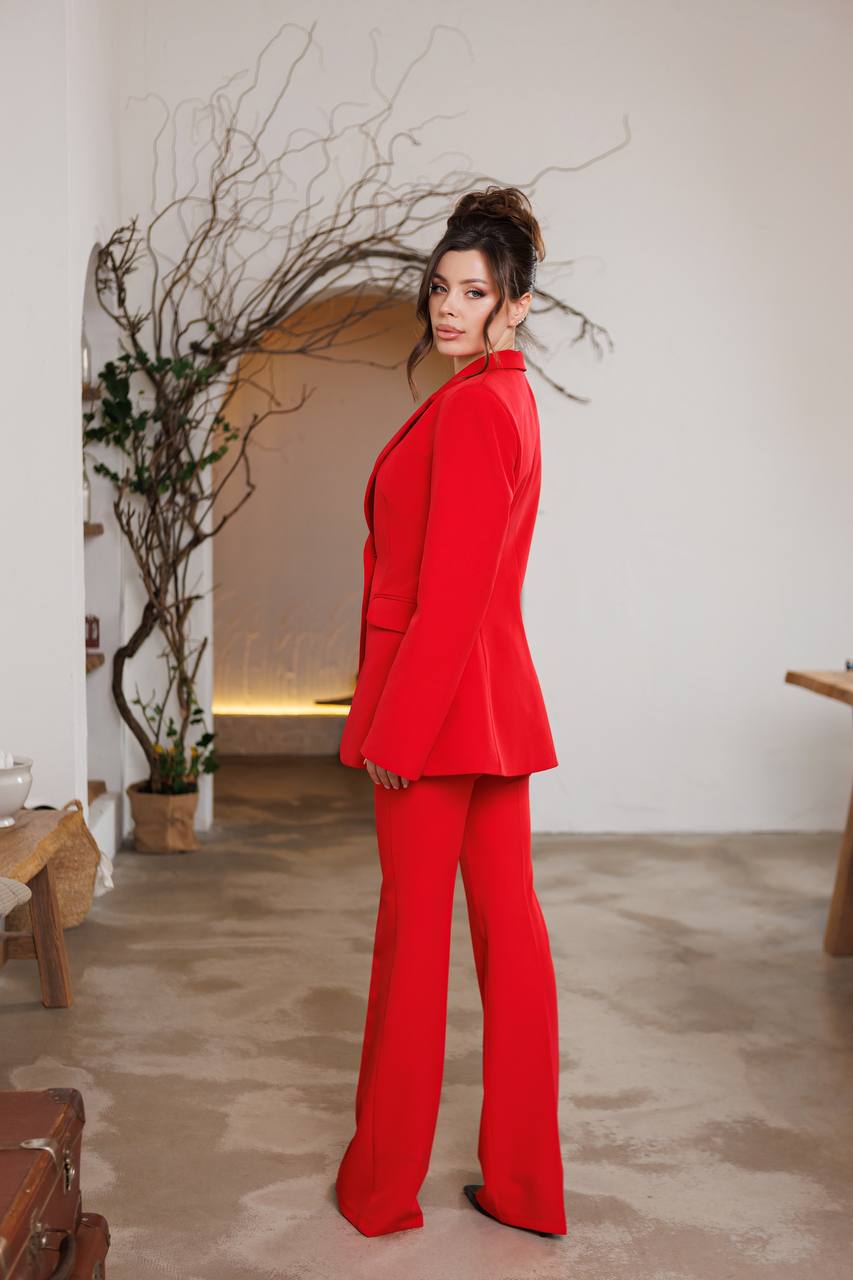 Woman in a red suit standing in a room with a plant and wooden furniture.