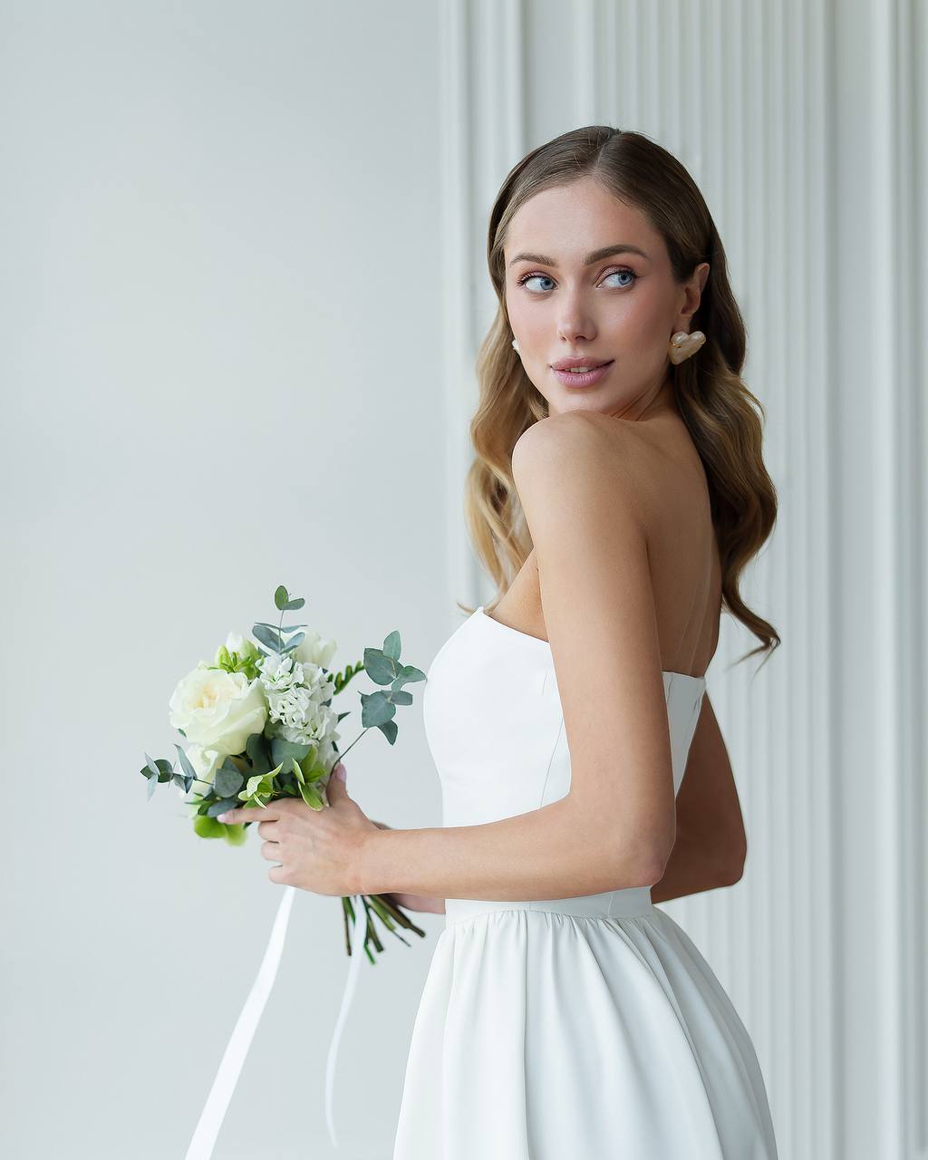 Woman in a white wedding dress holding a bouquet against a light gray background