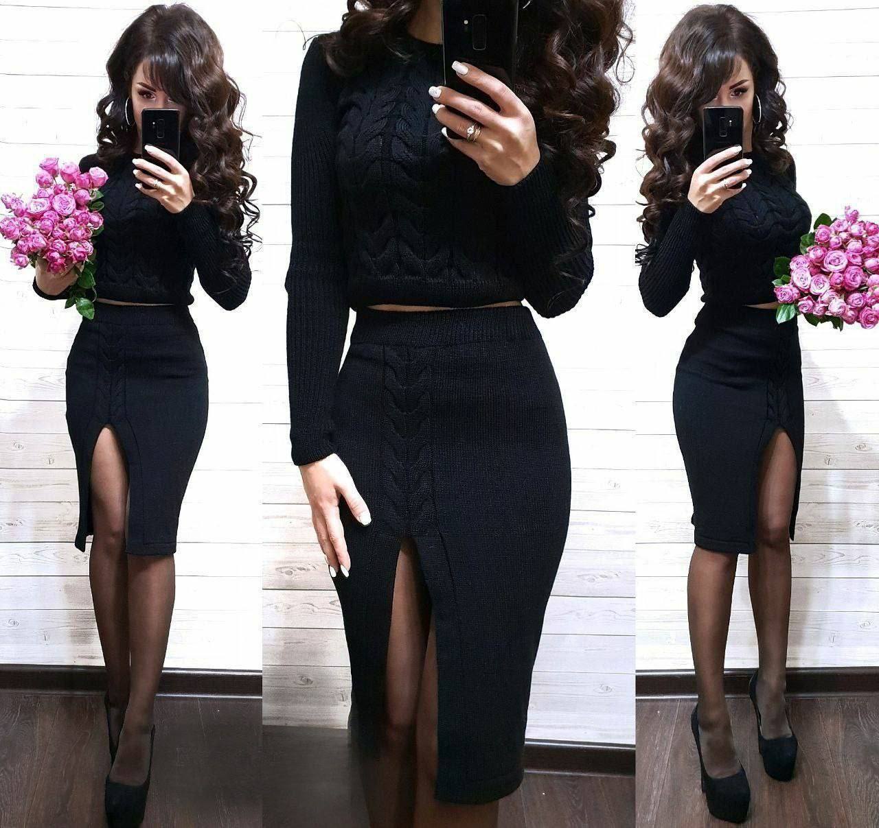 Woman wearing a black outfit with a high slit, holding pink flowers against a white wall.