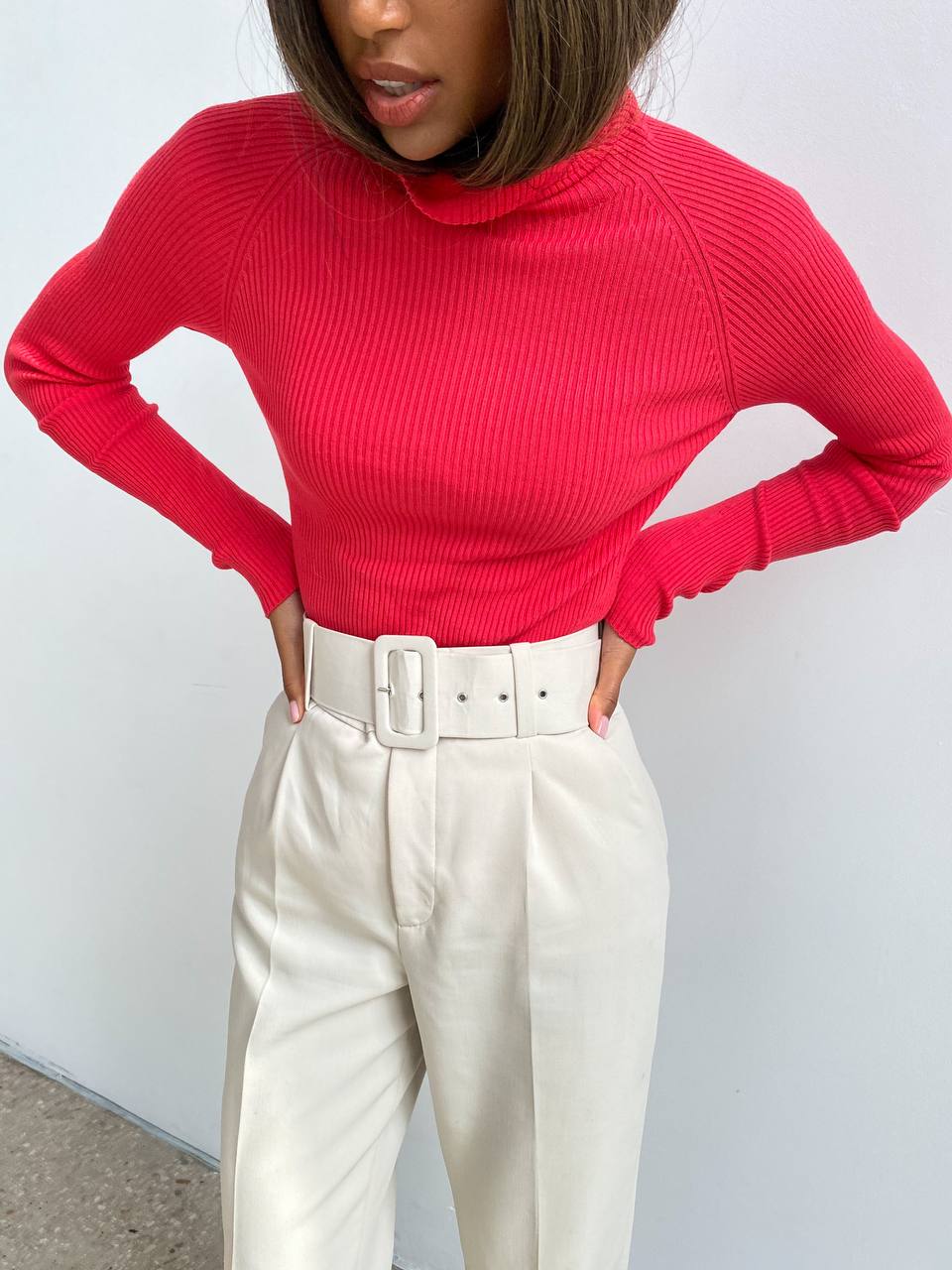 Person wearing a red ribbed sweater and white pants against a plain background