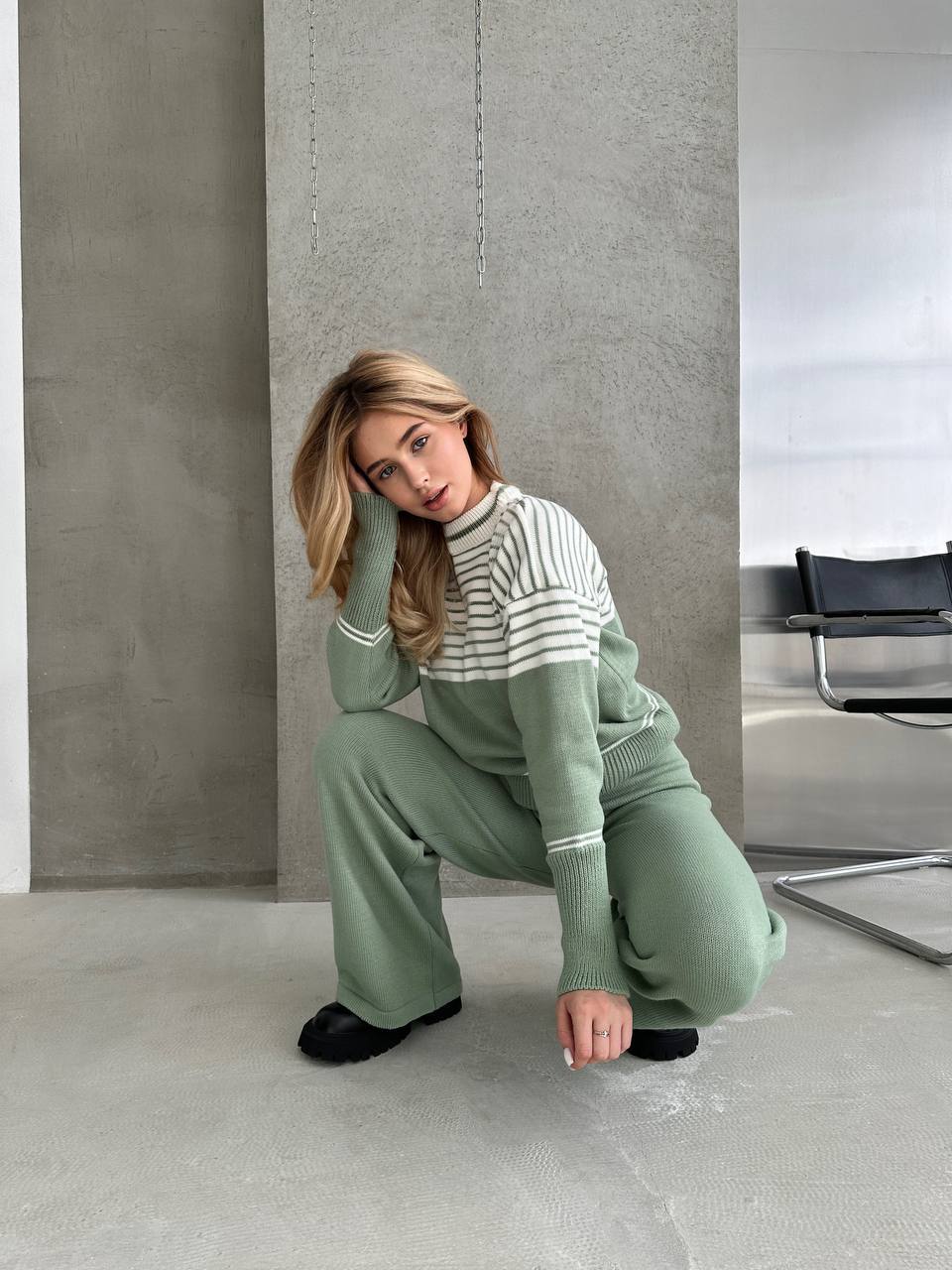 Woman in a green tracksuit and striped shirt posing in a modern room.