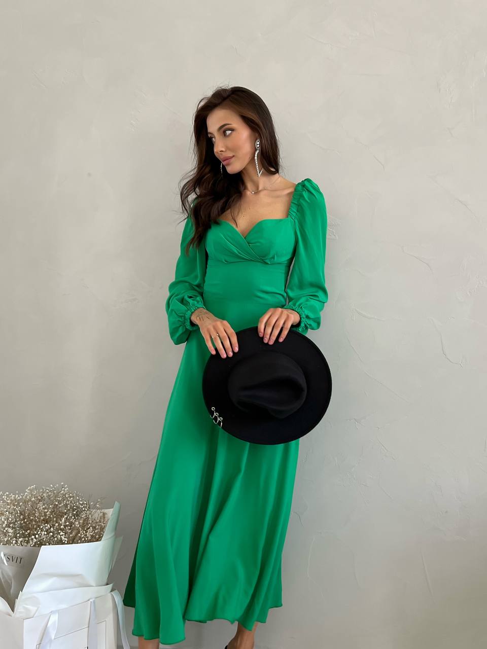Woman in a green dress holding a black hat against a neutral background
