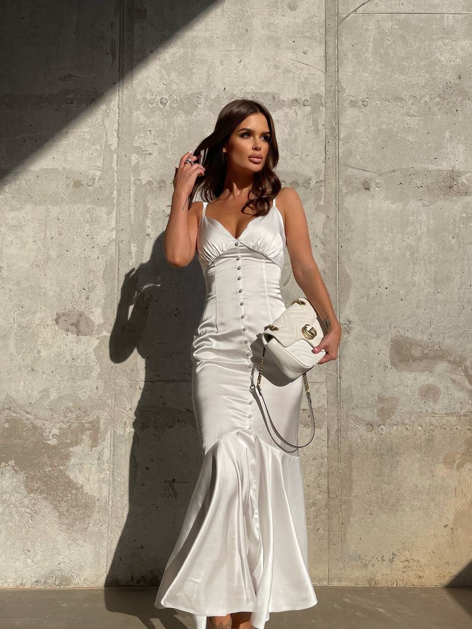 Woman in a white dress holding a white handbag against a concrete wall.