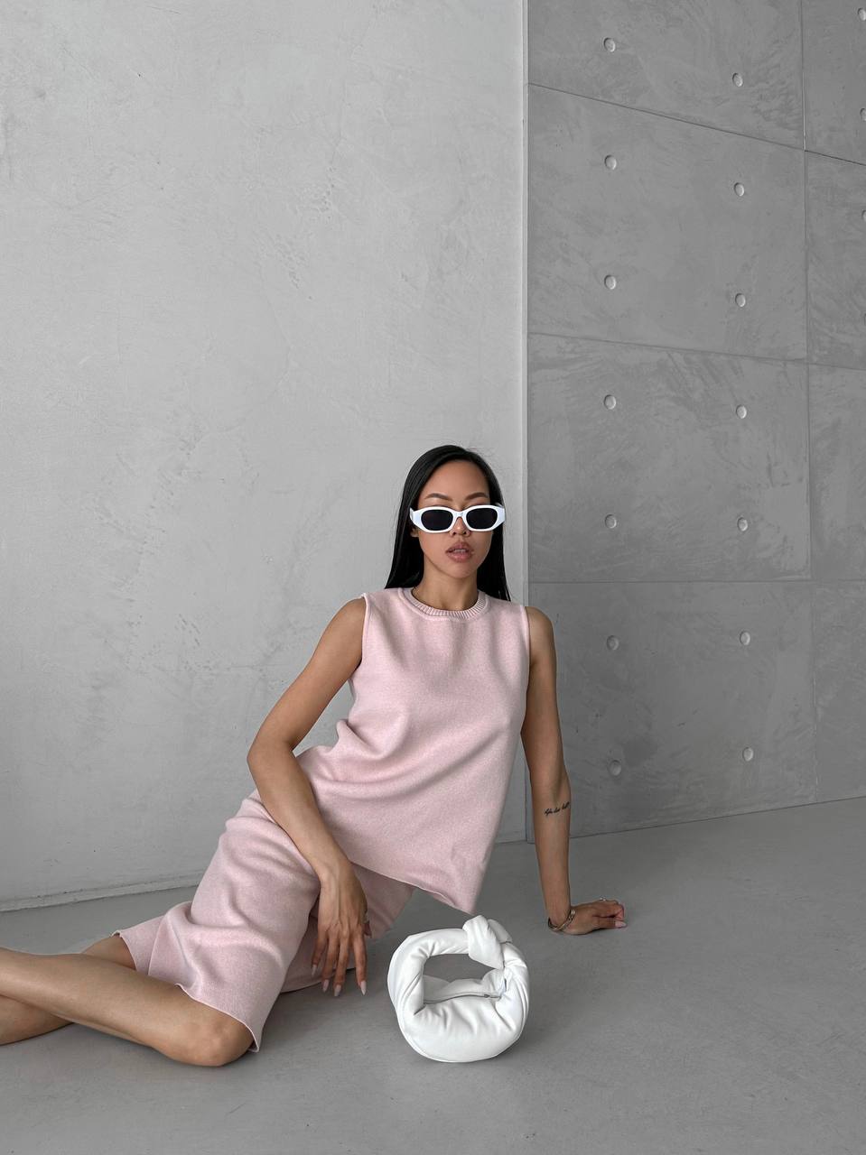 Woman in a pink dress sitting on the floor next to a white bag against a gray concrete wall.