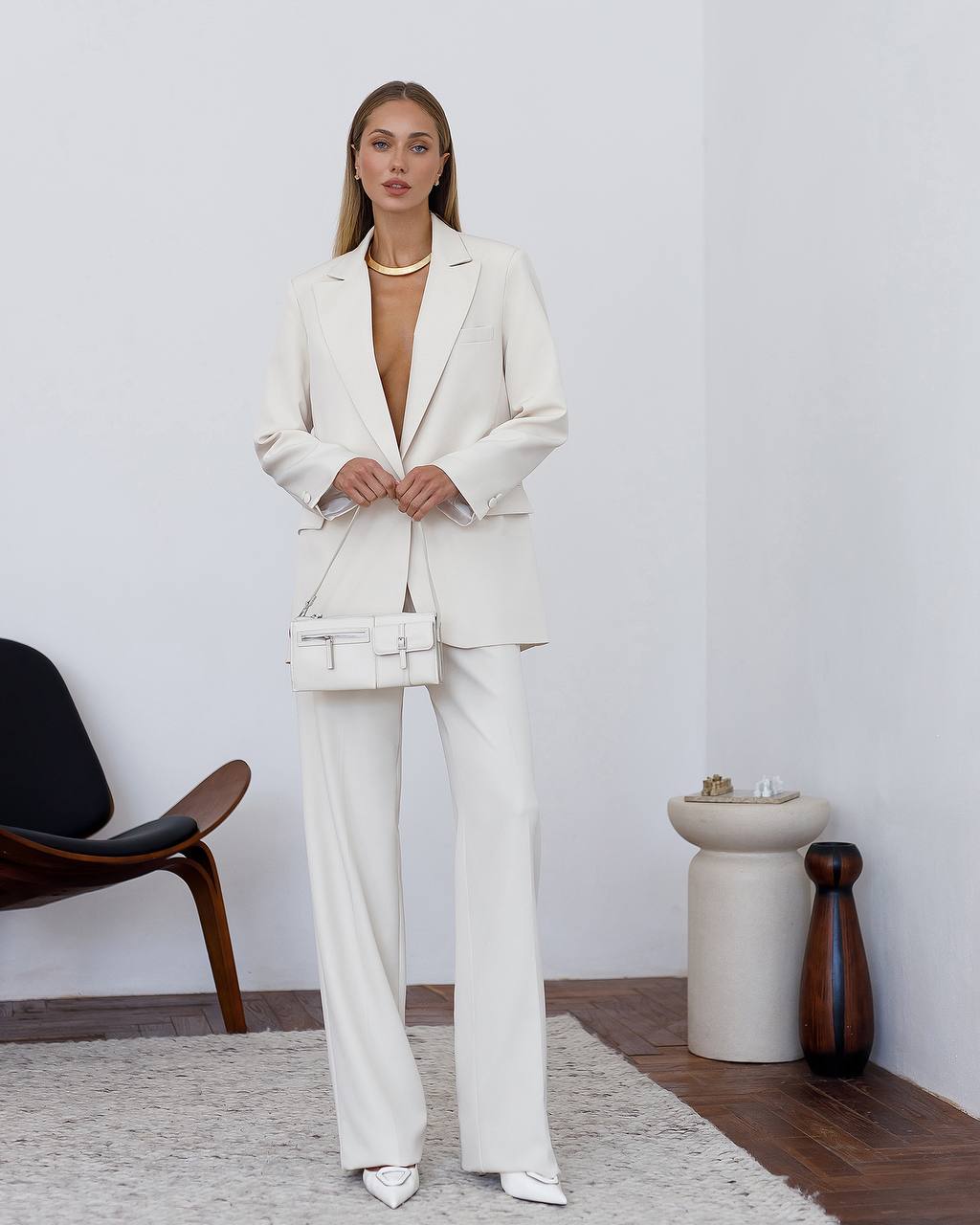 Woman wearing a white suit standing in a minimalistic room with a chair and table.