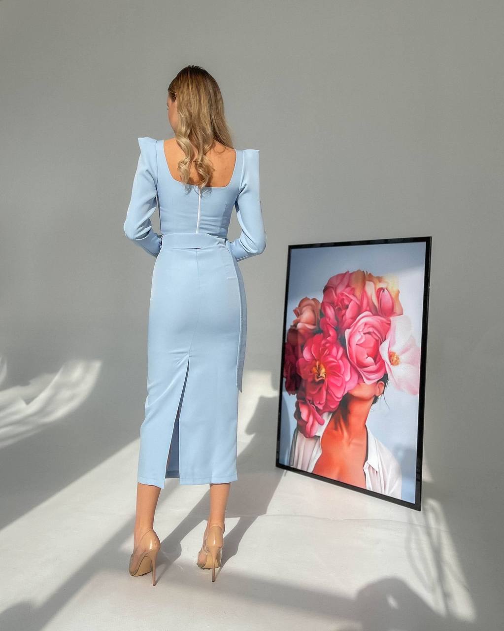 Woman in a light blue dress standing next to a framed artwork of a woman with flowers on her head.