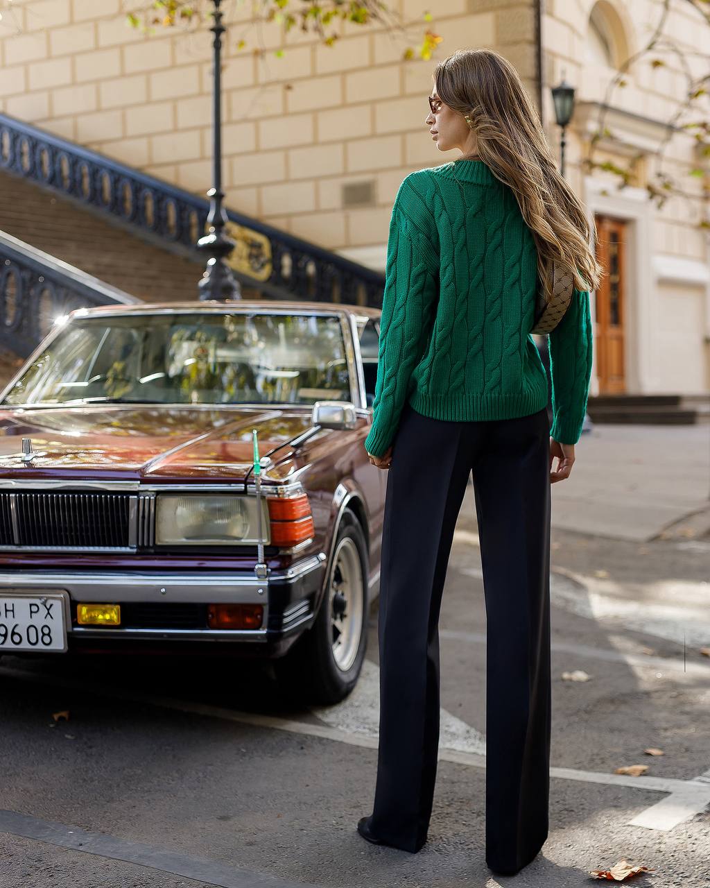 Woman in a green sweater and black pants standing next to a vintage car on a city street.