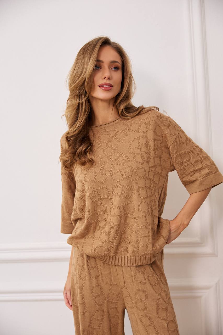 Woman wearing a brown textured sweater and pants against a white wall.