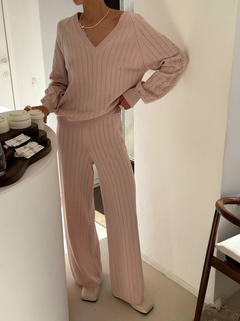 Person wearing a pink ribbed sweater and pants in a minimal indoor setting.