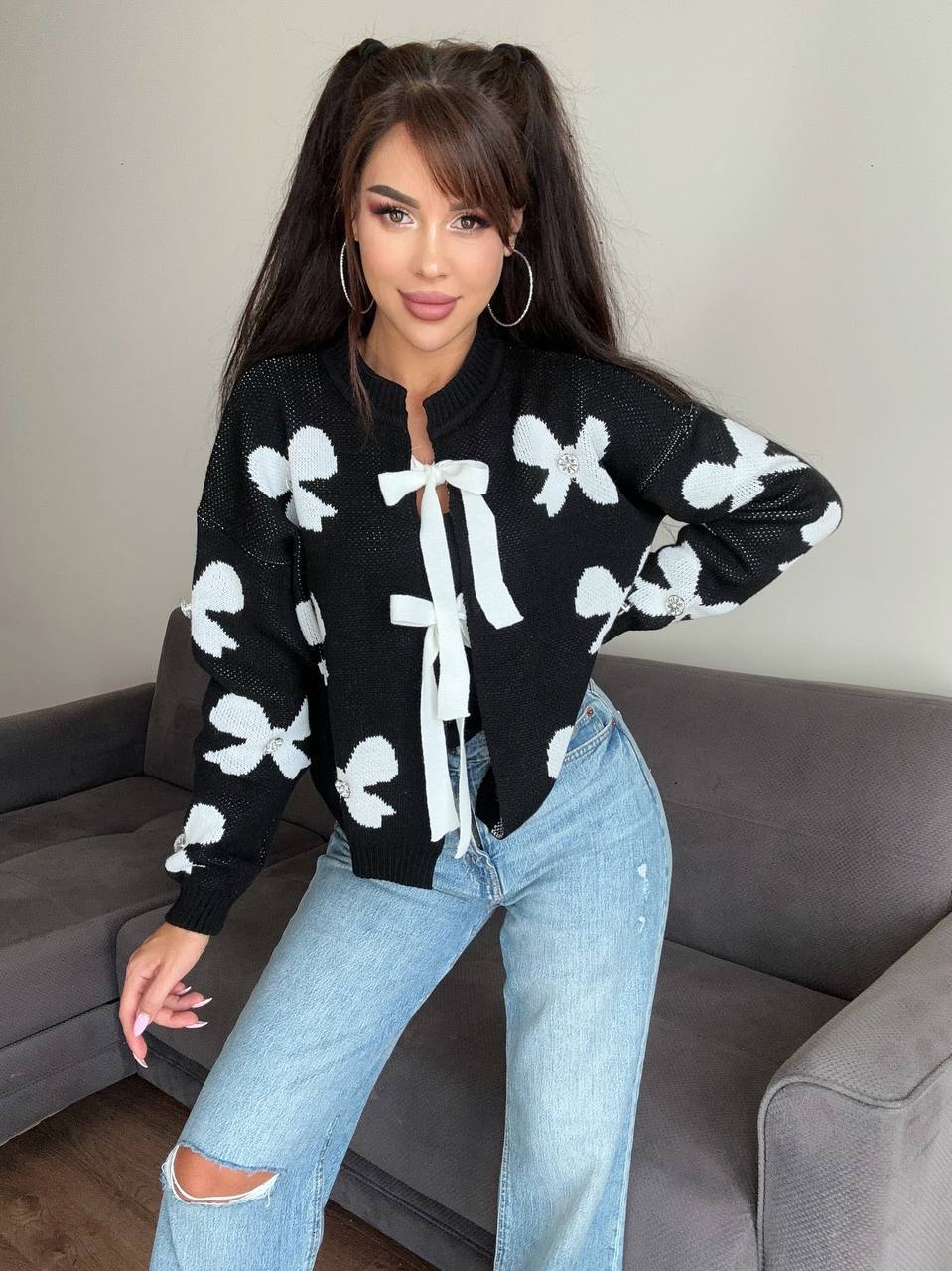 Woman wearing a black sweater with white bow patterns and blue jeans in a living room.