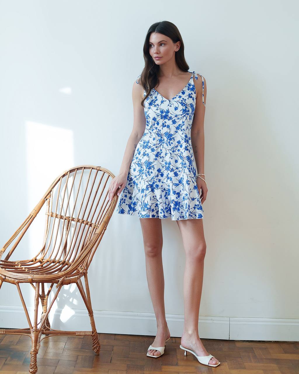 Woman in a blue floral dress standing next to a wicker chair against a white wall.