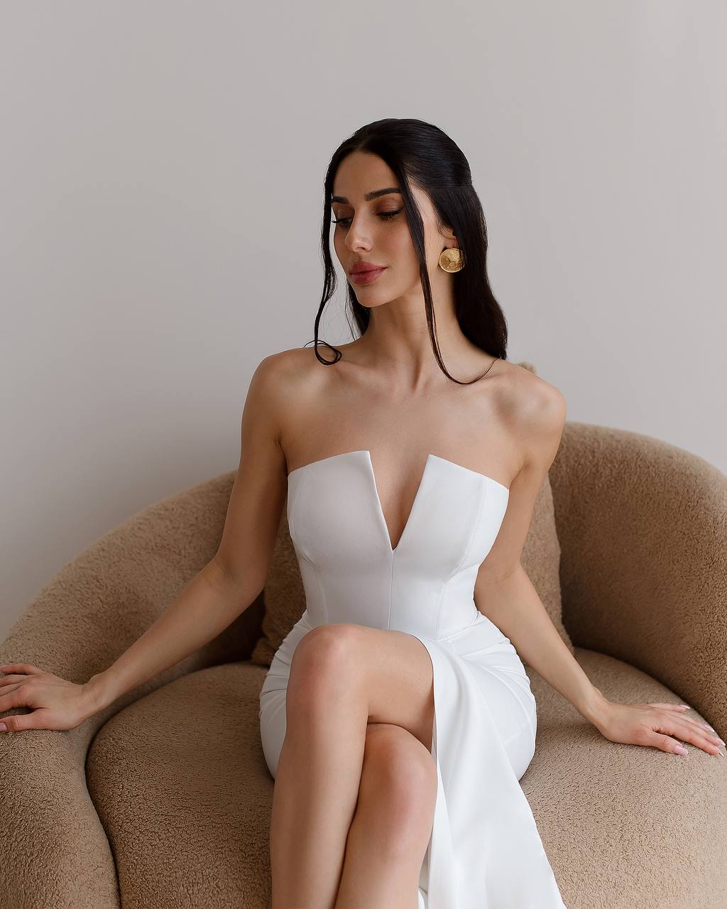 Woman in a white strapless dress sitting on a brown couch against a plain background