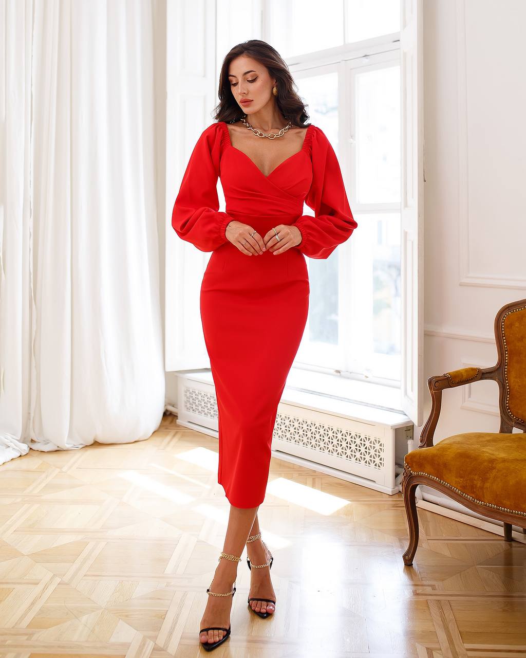 Woman wearing a red dress standing in a well-lit room with white curtains and a wooden floor.