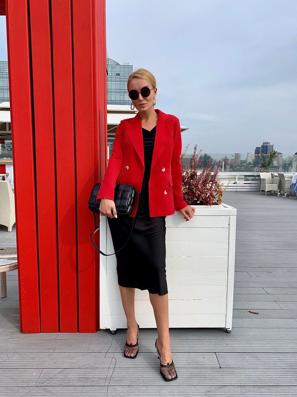 Woman in a red blazer and black skirt standing next to a red wall with a cityscape in the background.