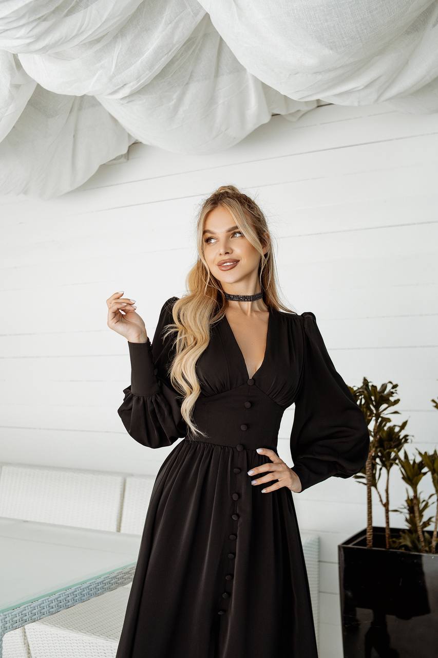 Woman in a black dress standing indoors with white walls and a plant in the background