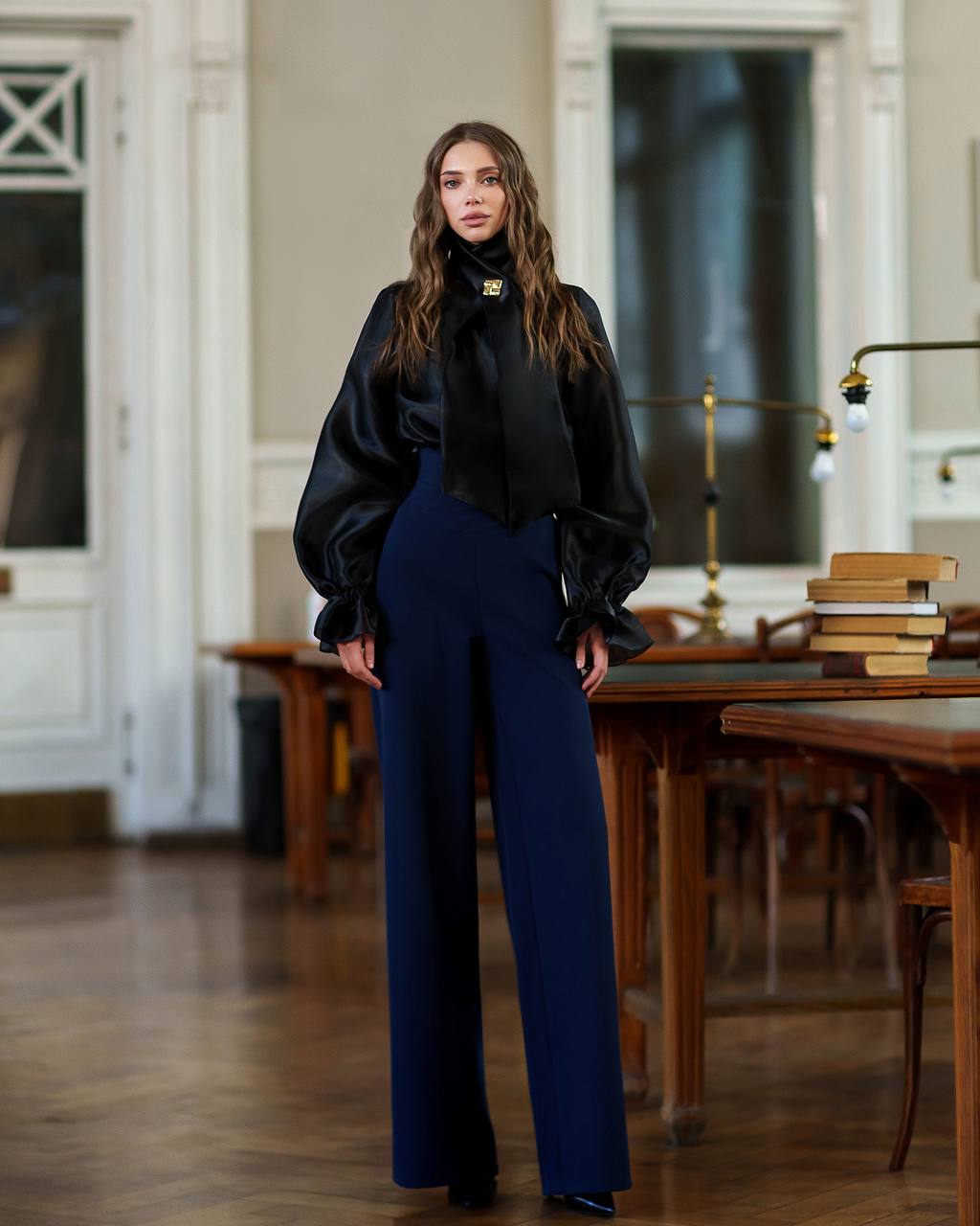 Woman in black top and blue pants standing in a room with wooden tables and books.