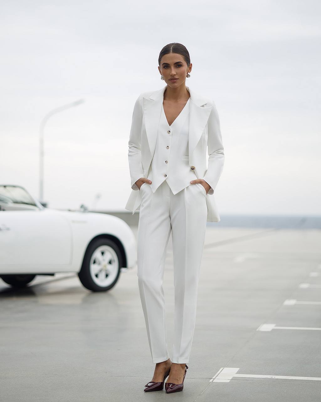 Woman in a white suit standing in front of a white car on a road.