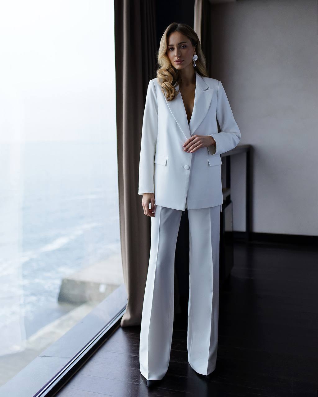 Woman in a white suit standing in a room with large windows.