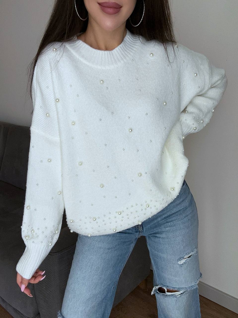 Person wearing a white sweater with pearl embellishments and blue jeans indoors.