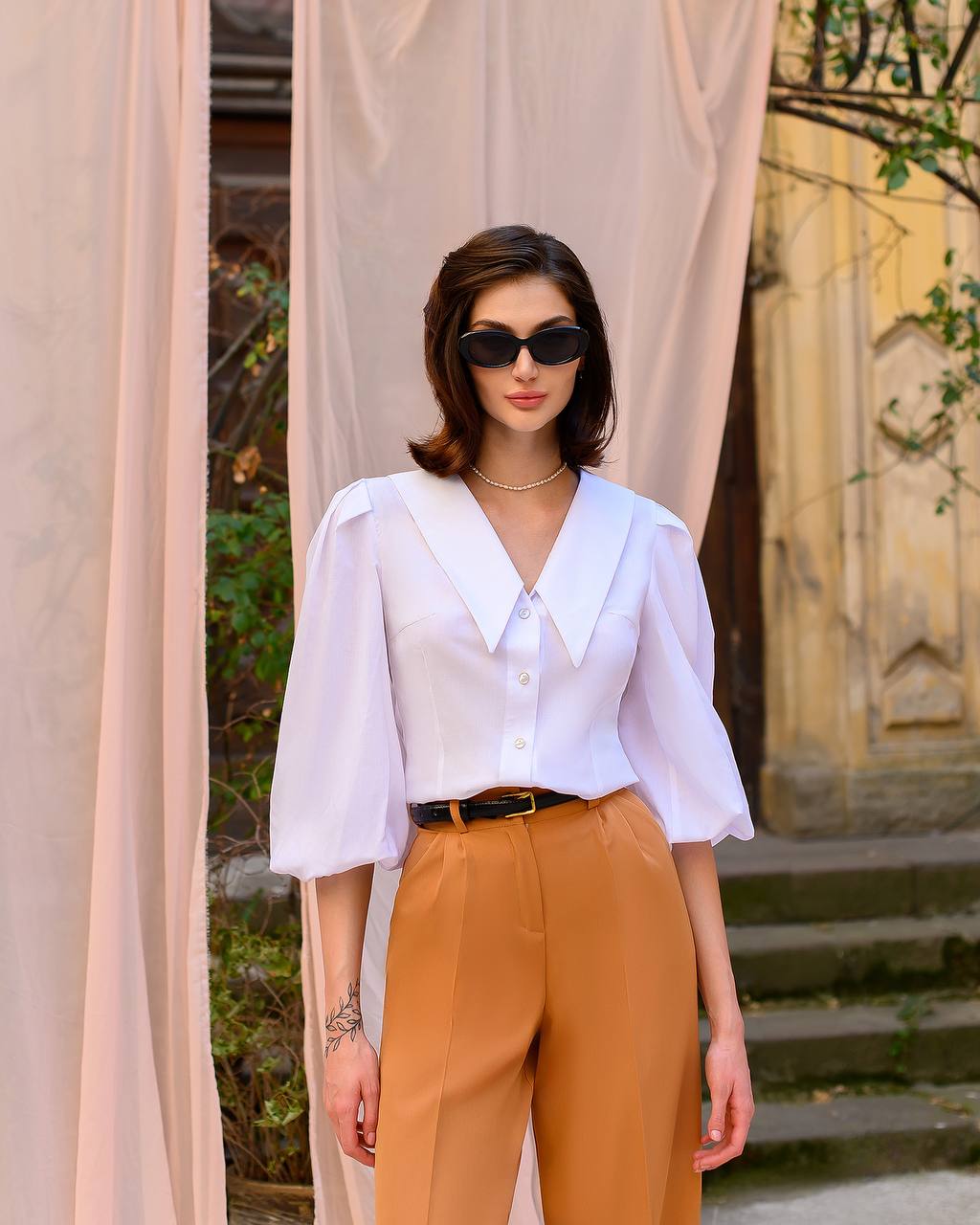 Woman wearing a white blouse and brown pants standing in front of beige curtains.