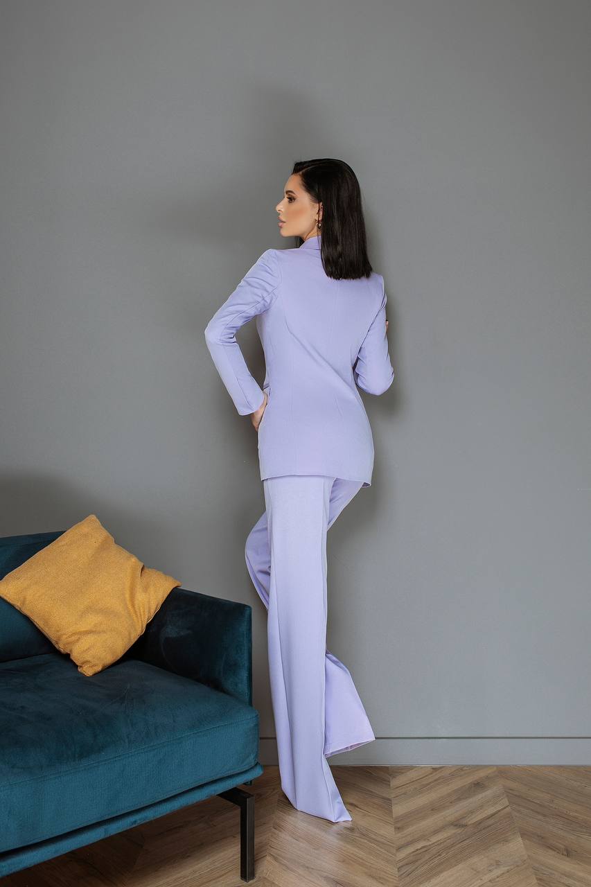 Woman in a light purple outfit standing in a room with a teal sofa and yellow pillow.